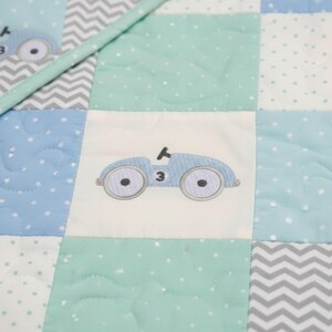 May include: A patchwork quilt featuring a repeating pattern of squares in shades of blue, mint green, and white. Each square has a different design, including a light blue embroidered car with the number 3, polka dots, and a chevron pattern. The quilt is quilted with a diamond pattern.