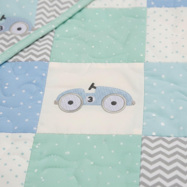 May include: A patchwork quilt featuring a repeating pattern of squares in shades of blue, mint green, and white. Each square has a different design, including a light blue embroidered car with the number 3, polka dots, and a chevron pattern. The quilt is quilted with a diamond pattern.