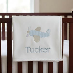 May include: A white baby blanket with a blue and tan embroidered airplane design. The name "Tucker" is embroidered in blue below the airplane. The blanket is draped over a dark brown crib.