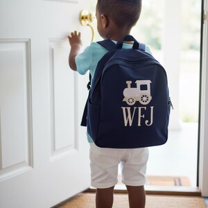 May include: A navy blue backpack with a white train design and the initials "WFJ" is worn by a child. The child is wearing a light blue shirt and white shorts, standing near a white door.