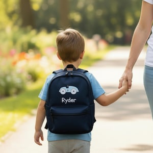 May include: A navy blue backpack with a light blue car design and the name "Ryder" in white. The backpack has a front pocket and a top handle. The child is wearing a light blue shirt and gray pants.