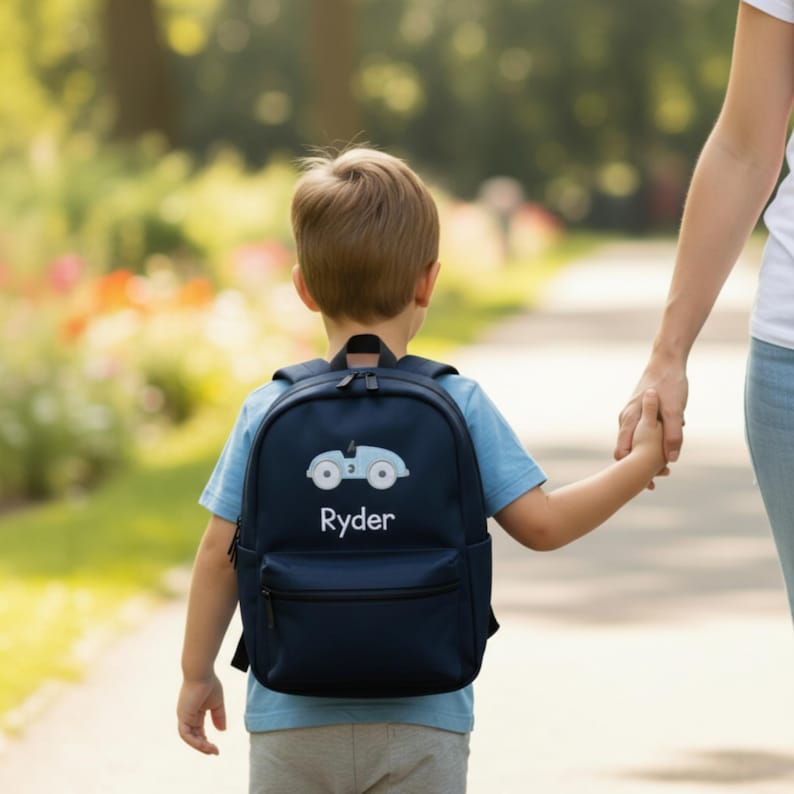 May include: A navy blue backpack with a light blue car design and the name "Ryder" in white. The backpack has a front pocket and a top handle. The child is wearing a light blue shirt and gray pants.