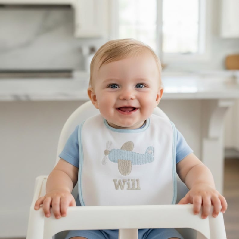 May include: A baby wearing a blue short-sleeved shirt and a white bib with a blue airplane design. The bib has a blue trim and the name "Will" embroidered in beige.
