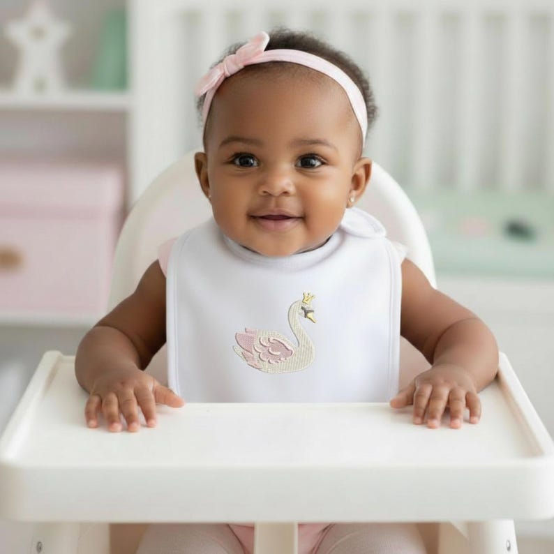 May include: A baby wearing a white bib with a pink swan design and a pink bow headband. The baby is sitting in a white high chair and smiling.