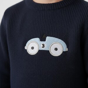 May include: Navy blue knit sweater featuring a light blue embroidered race car design. The car has white wheels with black centers and the number 3. The sweater has a ribbed crew neck and cuffs.