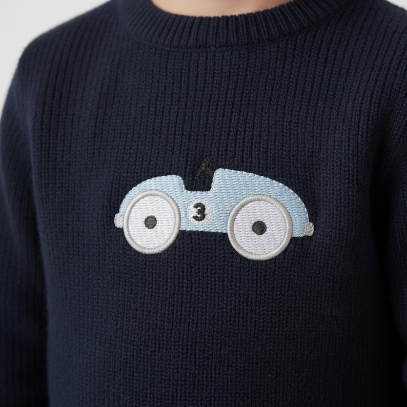 May include: Navy blue knit sweater featuring a light blue embroidered race car design. The car has white wheels with black centers and the number 3. The sweater has a ribbed crew neck and cuffs.