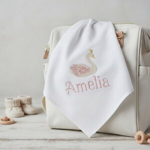 May include: White baby blanket with an embroidered swan design in pink and gold, with the name "Amelia" in pink script. The blanket is draped over a white diaper bag, with baby booties and a wooden toy in the background.