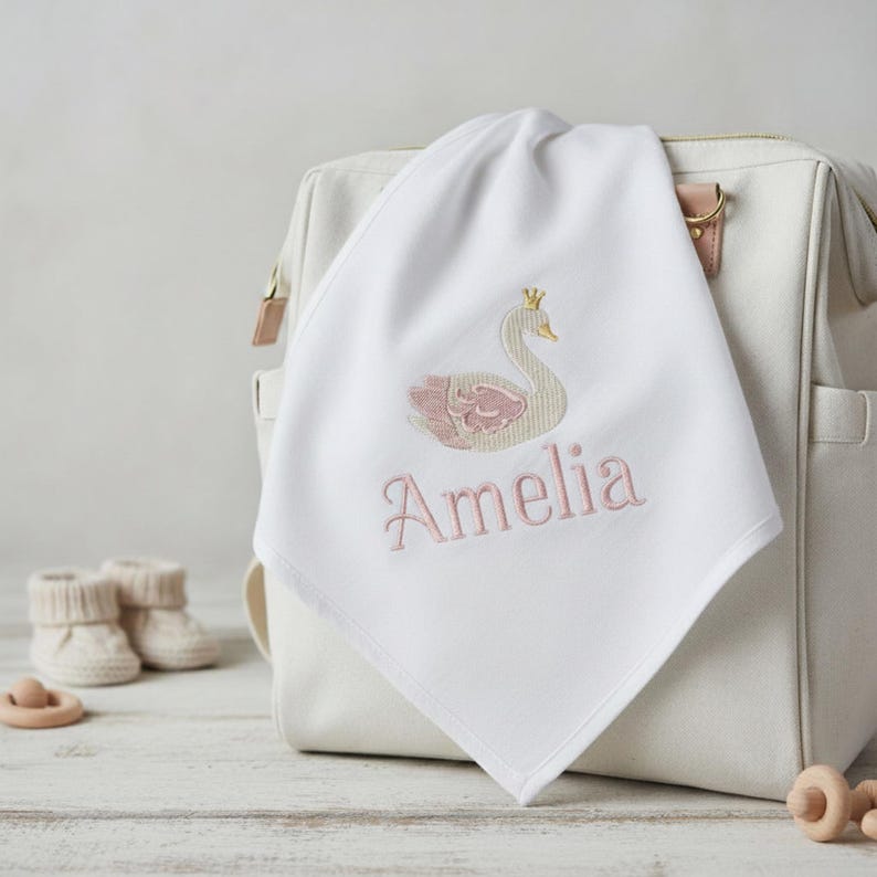 May include: White baby blanket with an embroidered swan design in pink and gold, with the name "Amelia" in pink script. The blanket is draped over a white diaper bag, with baby booties and a wooden toy in the background.