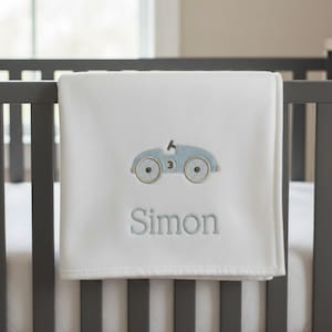 May include: White baby blanket with a light blue embroidered race car design and the name "Simon" in light blue script. The blanket is draped over a gray crib railing.