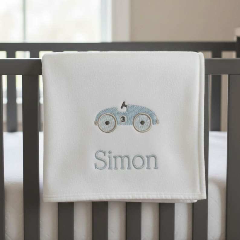May include: White baby blanket with a light blue embroidered race car design and the name "Simon" in light blue script. The blanket is draped over a gray crib railing.