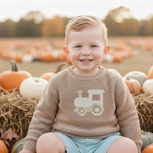 May include: A young child wearing a tan sweater with a light-colored train design, sitting among pumpkins and hay bales. The child is smiling, with a pumpkin patch in the background.