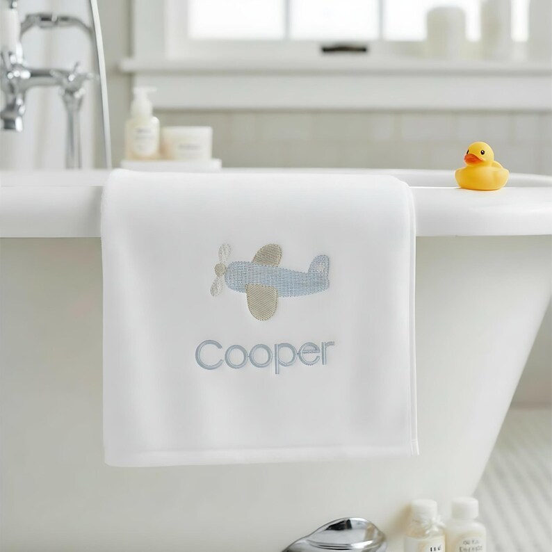 May include: White baby bath towel with an embroidered light blue and tan airplane design and the name "Cooper" in light blue. A yellow rubber duck sits on the edge of a white bathtub.