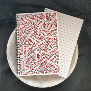 May include: A spiral-bound notebook with a cover featuring a collage of Diet Coke cans in red and white. A lined notepad rests beside it, both on a marble-patterned tray. The background is dark.