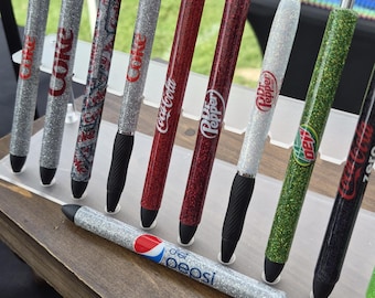 May include: A collection of glitter pens with various soda brand logos, including Coke, Coca-Cola, Dr. Pepper, and Diet Pepsi. The pens have silver, red, and green glitter barrels with black grips and silver accents. The pens are displayed on a wooden stand.