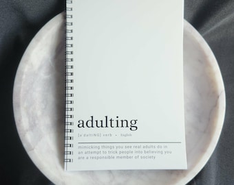 May include: A white spiral-bound notebook with the word "adulting" printed on the cover, along with a definition. The notebook rests on a marble-patterned tray, set against a dark gray background.
