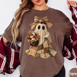 May include: An espresso-colored t-shirt featuring a graphic of a ghost wearing a burlap bow and holding a basket of pumpkins. The ghost has a patchwork-style skirt with various tan, brown, and green patterns. A red and white plaid shirt is layered over.