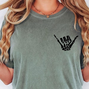 Pode incluir: Uma mulher vestindo uma camiseta verde musgo com um gr&aacute;fico de m&atilde;o de esqueleto preto "hang loose". A camiseta tem um caimento relaxado e mangas curtas.