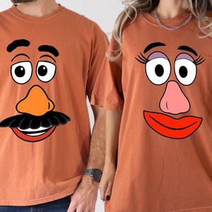 May include: Two rust-colored t-shirts, each featuring a cartoon face design. One shirt has a mustache, orange nose, and a smiling mouth. The other shirt has eyelashes, a pink nose, and red lips. The shirts are short-sleeved.