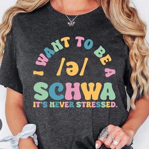 May include: Dark gray t-shirt with the text "I WANT TO BE A SCHWA. IT'S NEVER STRESSED." in colorful, retro-style lettering. The shirt is a heather dark gray color.