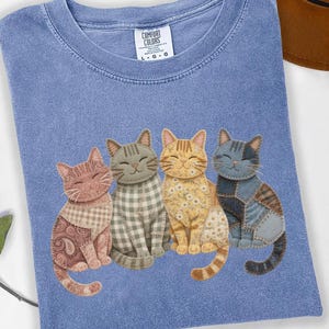 May include: A blue Comfort Colors t-shirt featuring four cartoon cats with unique patterns and colors. The cats are arranged in a row, each with a distinct design, including paisley, plaid, and patchwork. The shirt has a crew neck and a small tag.