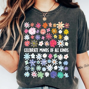 May include: Dark gray t-shirt featuring a colorful floral design. The shirt displays a grid of various flowers in shades of pink, orange, yellow, blue, and green. The text "CELEBRATE MINDS OF ALL KINDS" is printed in white.