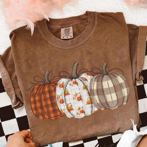 May include: Brown t-shirt featuring three decorative pumpkins with different patterns. The pumpkins include orange plaid, white floral, and brown plaid designs. The shirt has a Comfort Colors tag.