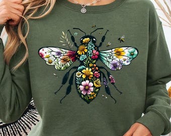 May include: Olive green sweatshirt featuring a large, detailed bee design. The bee's body is filled with colorful flowers, and its wings display a mix of floral patterns. The sweatshirt has long sleeves and a crew neck.