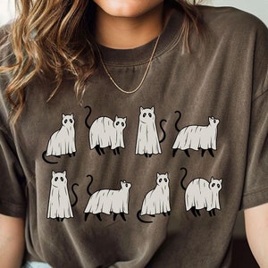 May include: A dark brown t-shirt with a graphic print of nine white ghost cats. The cats are in various poses, some are sitting, some are standing, and some are walking.