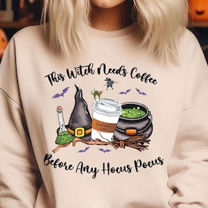 May include: A beige sweatshirt with a graphic print of a witch's hat, a coffee cup, a cauldron, and bats. The text on the sweatshirt reads "This Witch Needs Coffee Before Any Hocus Pocus".