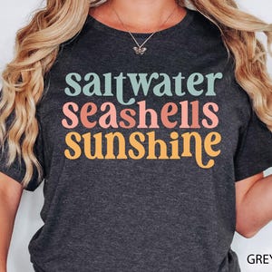 May include: Dark grey heather t-shirt featuring the words "saltwater seashells sunshine" in teal, coral, and yellow. The t-shirt has a crew neck.