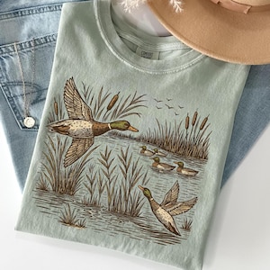 May include: A light green t-shirt featuring a detailed illustration of ducks in a marsh setting. The design includes flying ducks, reeds, and ducks swimming in the water. The shirt is paired with blue jeans and a tan hat.