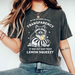 May include: A charcoal gray short-sleeve t-shirt featuring a white graphic of a raccoon holding a yellow lemon. The text reads: IN THE INTEREST OF FULL TRANSPARENCY IT WAS NOT EASY PEASY LEMON SQUEEZY. The design includes small star accents.