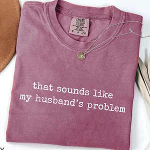 May include: Berry-colored t-shirt with the phrase "that sounds like my husband's problem" in white text. A silver necklace with a pendant is draped across the shirt. A brown hat and dried plant are in the background.