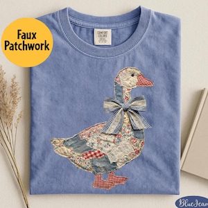 May include: A light blue t-shirt with a faux patchwork goose design. The goose features a patchwork of floral, denim, and gingham fabrics, with a blue and white checkered bow. The shirt has the text "Faux Patchwork" in a yellow circle.