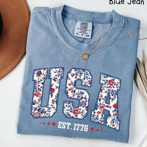 May include: Blue jean-colored t-shirt with the word "USA" in a floral pattern, and the text "EST. 1776". A gold necklace with a pendant is on the shirt. A brown hat is visible in the background.