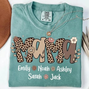 May include: A teal Comfort Colors t-shirt with the word "MAMA" in a mix of leopard print and tan letters, decorated with floral accents. Below the word are the names Emily, Noah, Ashley, Sarah, and Jack.