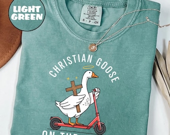 Può includere: T-shirt verde chiaro con la scritta "CHRISTIAN GOOSE ON THE LOOSE" e un'oca dei cartoni animati su uno scooter rosso, che tiene una croce. La maglietta ha un girocollo e maniche corte. Una collana d'argento è sulla maglietta.