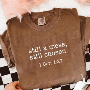 May include: A brown Comfort Colors t-shirt with the text "still a mess, still chosen. 1 Cor. 1:27" in white. The shirt is folded, with the sleeves rolled up. The background includes a black and white checkered pattern and pink decorative elements.