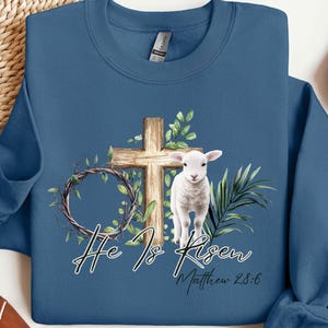 May include: A blue sweatshirt with a white lamb standing in front of a wooden cross. The cross is surrounded by green leaves and vines. The text "He Is Risen" is written in black cursive lettering below the cross. The text "Matthew 28:6" is written in black lettering below the cursive text.