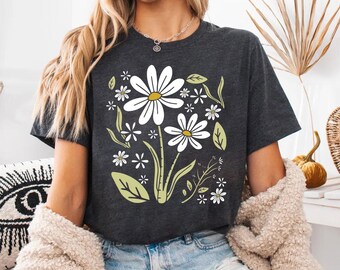 May include: A dark charcoal gray short-sleeve t-shirt featuring a central graphic print of white daisies with yellow centers, accompanied by light green leaves and stems. The design includes various sizes of flowers arranged in a cluster.