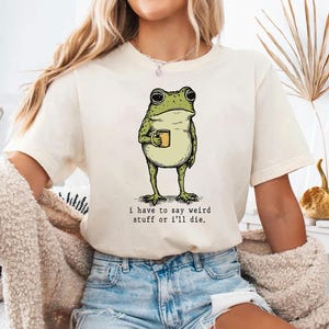 May include: Cream-colored t-shirt featuring a green frog holding a yellow mug. The frog illustration has a vintage style. The text "i have to say weird stuff or i'll die" is printed below the frog. The shirt is made of soft material.