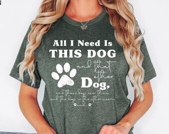 May include: A heather forest green short-sleeve t-shirt featuring white text that reads All I Need Is THIS DOG and that other Dog, and these dogs over there, and the dogs in the other room, and... with a white paw print graphic.
