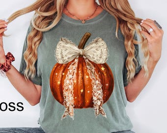 May include: A moss green short-sleeve t-shirt featuring a central graphic of a bright orange pumpkin adorned with a large, cream-colored lace bow. The pumpkin design includes small gold star accents and long, dangling lace ribbons extending downward.