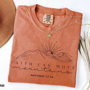 May include: A terracotta-colored short-sleeve t-shirt featuring a black line-art graphic of mountains and a rising sun. The text reads FAITH CAN MOVE mountains MATTHEW 17:20. The shirt has a crew neckline and a Comfort Colors brand label inside.