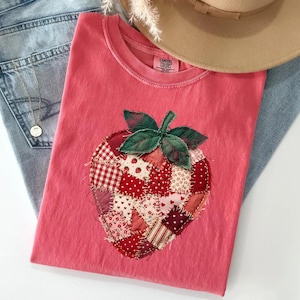 May include: A pink Comfort Colors t-shirt featuring a patchwork strawberry design. The strawberry is made of various red and white patterned fabrics with green leaves. A tan hat and blue jeans are also in the image.