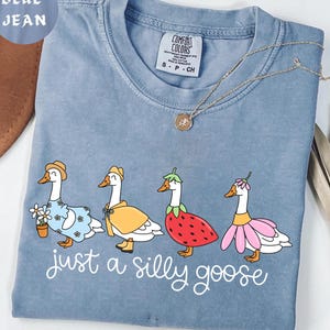 Puede incluir: Camiseta azul vaquero Comfort Colors con un dise&ntilde;o caprichoso de cuatro gansos vestidos con varios atuendos. La frase "just a silly goose" est&aacute; escrita en blanco. Un collar dorado est&aacute; sobre la camiseta. Talla S-P-CH.