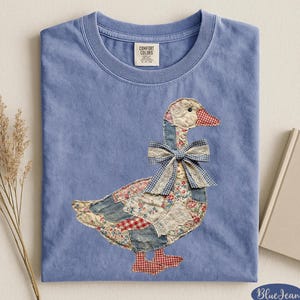May include: A blue Comfort Colors t-shirt featuring a patchwork goose design. The goose has a bow and is made of various fabrics, including floral and gingham patterns. The shirt has a crew neck and a tag that reads "Comfort Colors".
