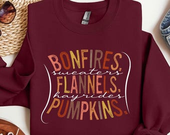May include: A maroon crewneck sweatshirt featuring a graphic print with the text BONFIRES, sweaters, FLANNELS, hayrides, PUMPKINS. The lettering uses a warm color palette of orange, yellow, and brown tones, arranged in a stacked, stylized layout.