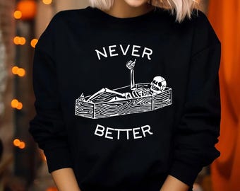 May include: A black long-sleeve sweatshirt featuring a white graphic print of a skeleton lying in a wooden coffin with a thumb up. The text NEVER is printed above the coffin and the text BETTER is printed below the coffin in white lettering.