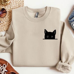 May include: A beige sweatshirt with a black cat peeking out from behind a pocket. The cat's eyes are white.